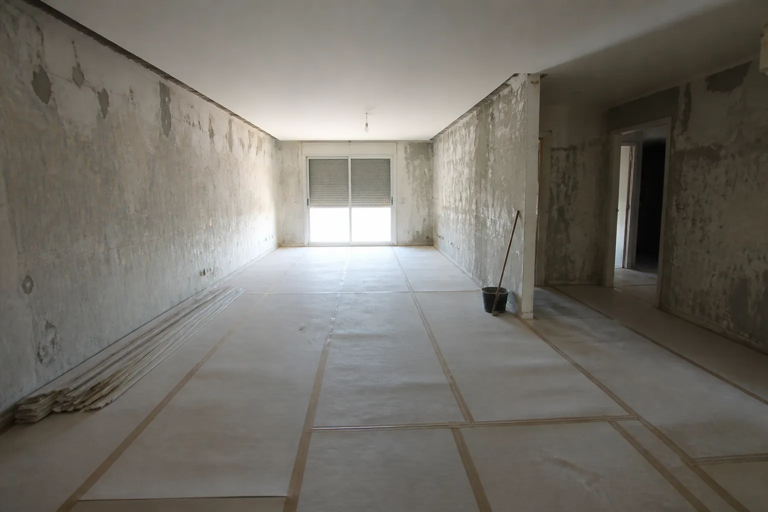Pièce principale d’un appartement entièrement vidé en cours de travaux, sol couvert de film et de cartons, murs mis à nu, volets à demi fermés