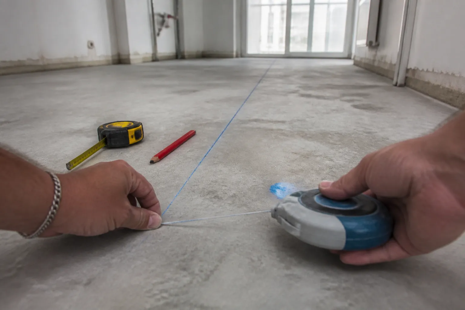 Deux mains d’artisan tendant un cordeau traceur au ras du sol d’un appartement vide pour marquer l’implantation d’une cloison